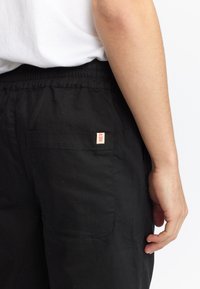 Mustat puuvillahousut, joissa on joustava vyötärönauha ja takaosa tasku, jossa on punainen merkkilappu. Kangas on sileä ja siinä on mattapintainen viimeistely.