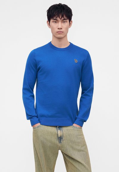 Jeune homme portant un pull à manches longues bleu vif avec un petit logo brodé sur la poitrine et un jean clair, les mains dans les poches.