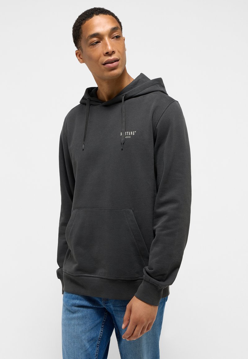 Sudadera de algodón gris con un bolsillo frontal, capucha con cordón y un pequeño logo en el pecho. Lleva puestas unas jeans de mezclilla azules. Textura suave.