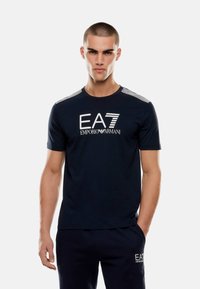 Mörkblå bomullströja med en vit EA7-logotyp, randiga detaljer på axlarna och en klassisk rundhalsdesign.