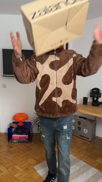 Smeđa jakna od umjetnog krzna s svijetlosmešanim apstraktnim uzorkom, zatvarač na zip, i izgužvane traperice. Kartonska kutija se drži u zraku.