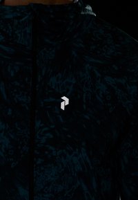 Veste à motifs noirs avec fermeture éclair, tissu texturé et un logo réfléchissant sur la partie supérieure gauche.