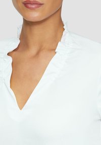 Weiße Bluse mit Rüschensaum, V-Ausschnitt und glatter Stoffstruktur. Nahaufnahme, die die Details des Ausschnitts hervorhebt.
