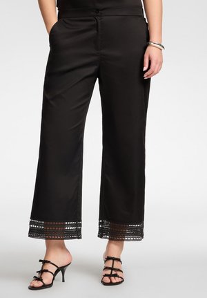 Mujer que lleva pantalones negros recortados con encaje en el dobladillo y sandalias negras de tacón con lazos, de pie con una mano en el bolsillo.