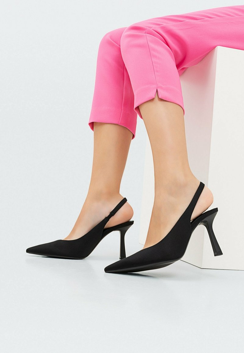 Jambes en pantalon court rose portant des escarpins slingback noirs à bout pointu avec talons aiguilles, assises sur un bloc blanc.