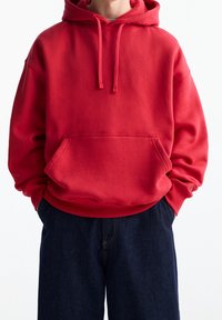 Czerwona bluza z kapturem, wyposażona w kieszeń kangurką, z prążkowanymi mankietami i dołem. W zestawie z ciemnymi dżinsami. Gładka faktura, casualowy design.