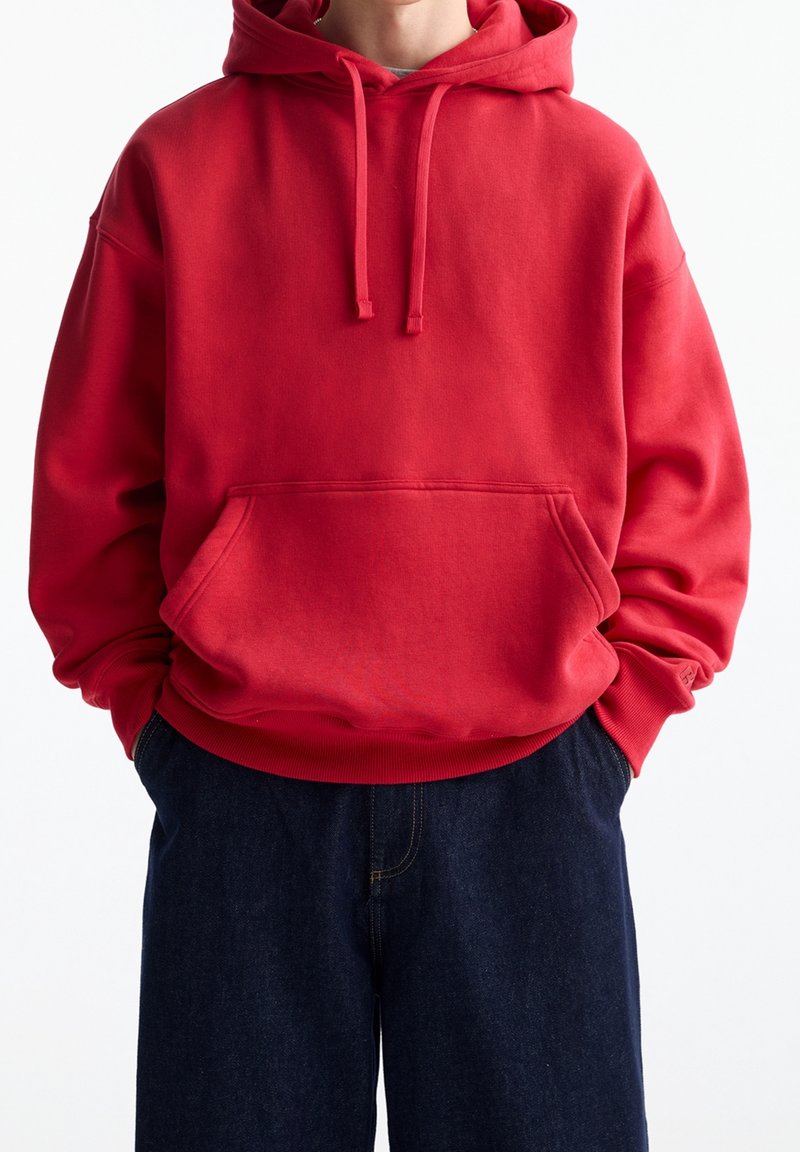 Czerwona bluza z kapturem, wyposażona w kieszeń kangurką, z prążkowanymi mankietami i dołem. W zestawie z ciemnymi dżinsami. Gładka faktura, casualowy design.