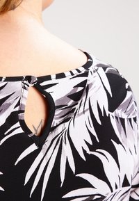 Haut noir avec motif de feuilles blanches et grises, présentant une découpe en forme de trou de serrure au niveau de l'encolure arrière et une finition en tissu texturé.