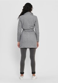 Cappotto grigio in misto lana con vita a cintura, maniche lunghe e colletto a risvolto, abbinato a pantaloni grigi aderenti e sneakers bianche.