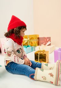 Rote, gerippte Strickmütze und Schal, cremefarbenes Langarmshirt mit festlichen Motiven, blaue Jeans und cremefarbene Regenstiefel mit Muster und festlichen Details.