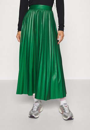 Personne portant une longue jupe plissée verte, un haut noir à manches longues, des chaussettes blanches avec des rayures roses et des chaussures de course argentées.