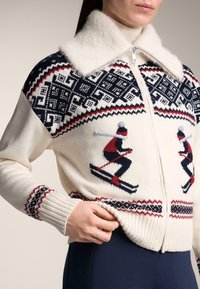 Sweter dziany o kremowej podstawie z granatowymi i czerwonymi wzorami geometrycznymi, przedstawiającymi postacie narciarzy. Miękki kołnierz z futerka; ściągacze na mankietach i dole. Zapięcie na zamek.