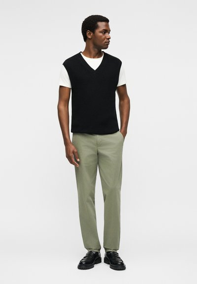 Άνδρας που στέκεται με τα χέρια στις τσέπες, φοράει μαύρο αμάνικο πουλόβερ με V-λαιμόκοψη πάνω από λευκό T-shirt, ανοιχτοπράσινο παντελόνι και μαύρα παπούτσια.