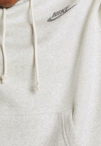 Ljusgrå hoodie med texturerat tyg, framficka och justerbara dragsnören. Har en diskret Nike-logotyp i mörkare färger.