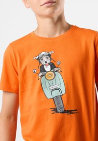 Orange bomulds t-shirt med et tegneseriehund, der bærer briller og kører på en lyseblå scooter, med en trykt tekstlabel "DENIM FACTORY."