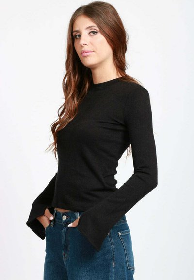 Donna con lunghi capelli castani, che indossa un top nero aderente a maniche lunghe con polsini svasati e jeans blu a vita alta, in piedi contro uno sfondo bianco.
