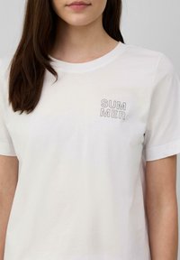 T-shirt en coton blanc avec un col rond et des manches courtes, arborant "ÉTÉ" imprimé en gris clair sur la partie supérieure gauche de la poitrine.