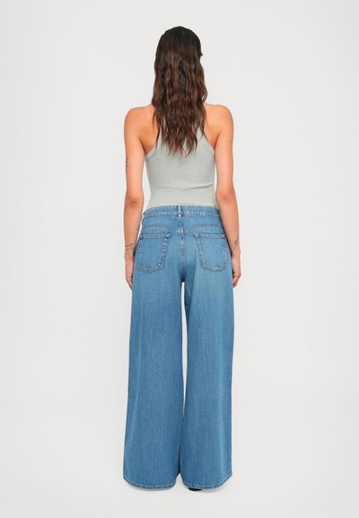Woman with long wavy hair wearing a gray sleeveless top and high-waisted wide-leg blue jeans, standing facing away against a plain background.