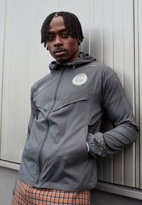 Chaqueta gris con cremallera y capucha, que presenta un logo circular en el pecho, tejido texturizado y puños elásticos. Se lleva con pantalones de cuadros.