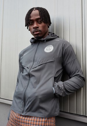 Grå zip-up jakke med hætte, prydet med et cirkulært logo på brystet, struktureret stof og elastiske manchetter. Bæres med ternede bukser.