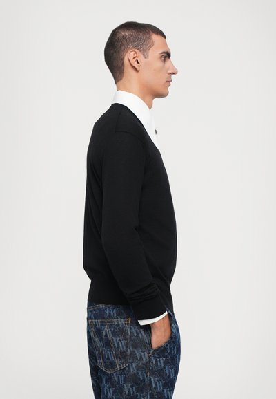 Black sweater with a crew neck, over a white collared shirt. Model stands sideways, wearing patterned blue jeans with visible pockets.