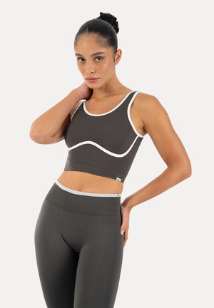 Kobieta nosząca ciemnoszary stanik sportowy i legginsy z białym wykończeniem, stojąca z jedną ręką na szyi, a drugą na biodrze.