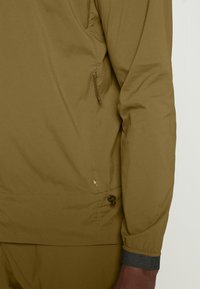 Gros plan d'une personne portant une veste vert olive avec une poche latérale zippée et un cordon de serrage réglable à l'ourlet.
