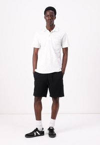 Hvit kortermet polo-shirt med brystlomme, kombinert med svarte shorts med snor. Modellen har på seg svarte joggesko med hvite detaljer.