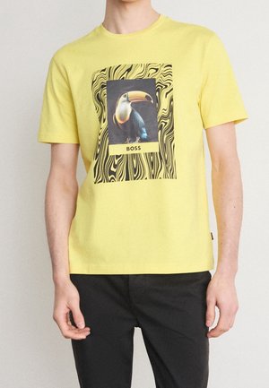 T-shirt en coton jaune avec un motif de toucan intégré dans un motif ondulé noir et blanc, avec une étiquette rectangulaire "BOSS" en bas.