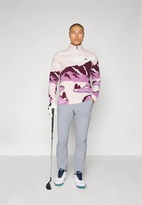 Uomo che indossa un pullover da golf rosa con motivo montano, pantaloni grigio chiaro, scarpe da golf bianche e un guanto, mentre tiene una mazza da golf.
