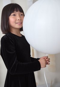 Ein Mädchen in einem schwarzen Samtkleid hält einen großen weißen Luftballon. Der Ballon ist rund, glatt und aufgeblasen, mit einem dünnen Band befestigt.