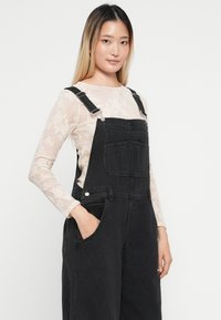 Črne denim naramnice z nastavljivimi trakovi, sprednjim žepom in stranskima žepoma, oblečene čez svetlo čipkast dolg rokav. Gladka tekstura.