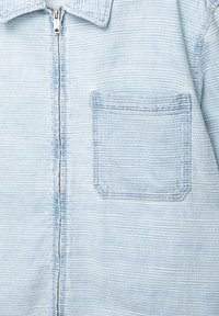 Svetle modrá džínová bunda s texturovaným povrchem, vybavená zipovým zapínáním a náprsní kapsou. Látka má jemné horizontální pruhy.