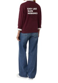 Bordeaux sweatshirt met witte tekst "EASY LIKE SLOW MORNINGS," gecombineerd met een wijde blauwe jeans en witte sneakers. Zachte stof, relaxte pasvorm.