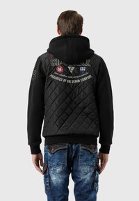 Schwarze gesteppte Jacke mit Kapuze, mit gestickten Logos und Akzenten, kombiniert mit blauen Denim-Cargohosen mit Taschen und Nähdetails.