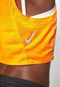 Gros plan d'une personne portant un haut de sport orange vif de Nike par-dessus un soutien-gorge de sport blanc et un short noir.