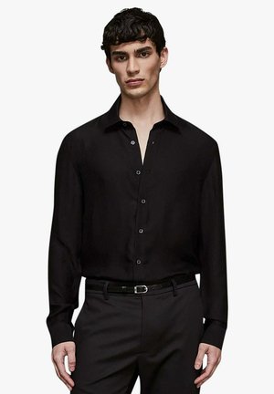 Giovane uomo che indossa una camicia nera a maniche lunghe con bottoni, infilata in pantaloni neri con cintura nera, in piedi contro uno sfondo bianco.