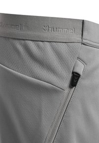 Grijze sportbroek in close-up met gestructureerde tailleband met "hummel" logo en een zijzak met rits en zwarte trekker.