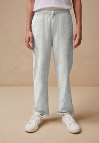 Pantalon de jogging gris clair texturé avec une taille élastique et un cordon de serrage. Associé à des baskets blanches sur un fond neutre.