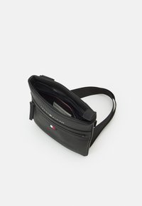Black, textured leather crossbody bag with a zipped main compartment and front pocket. Features a logo and red, white, and blue accent.