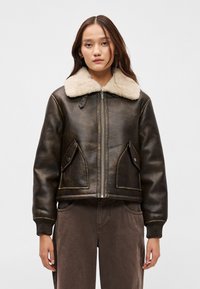 Smeđi kožni bomber jakna s krem boje umjetnim krznom ovratnikom, zip zatvaračem sprijeda i dva džepa s preklopima. Riblje manžetne i donji rub, teksturirani finiš.