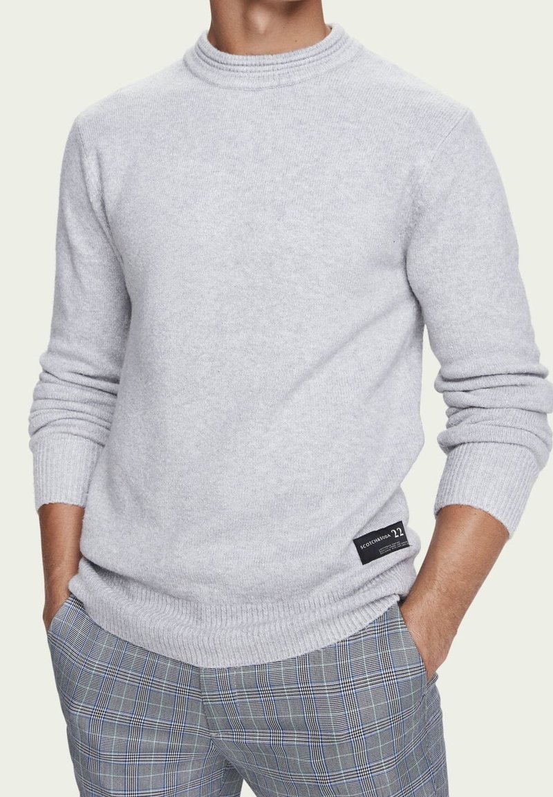 Pull en maille gris avec un col rond côtelé, des manches longues et un large ourlet ; présente un patch logo en bas à gauche.