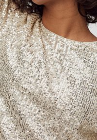 Haut beige présentant une surface texturée embellie de sequins disposés de manière uniforme, créant un effet scintillant sur le tissu.