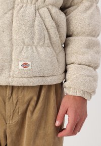 Beige pufferjacka med textilstruktur och elastiska muddar, med en framficka och Dickies-logotyp ovanför ett par kordbyxor.