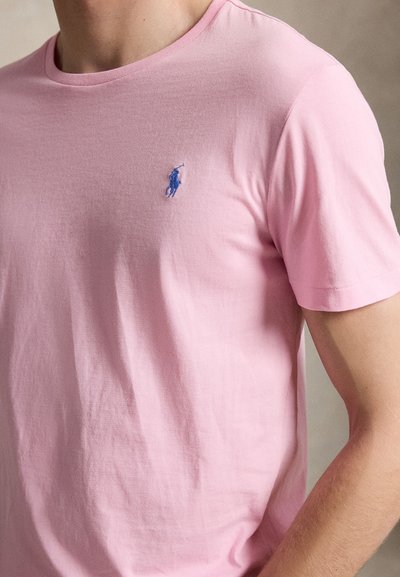 Mand iført en lyserød T-shirt med korte ærmer og en lille blå broderet polospiller-logo på venstre bryst.