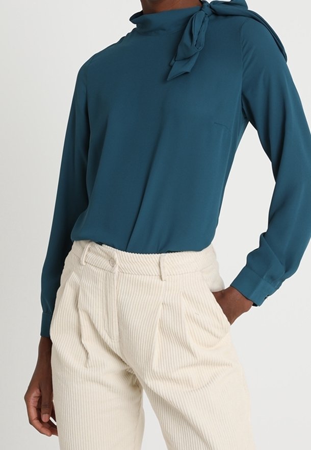 Teal blouse met een halsbandje, lange mouwen en een minimalistisch ontwerp, gecombineerd met crèmekleurige corduroy broeken met verticale strepen.