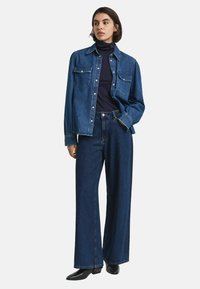 Vrouw in een donkerblauwe coltrui, losse spijkerbloes, wijde donkerblauwe jeans en zwarte puntige laarzen, staande met één hand in haar zak.