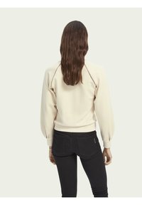 Felpa beige chiaro con maniche a sbuffo, polsini a coste e cuciture decorative lungo le cuciture delle spalle, abbinata a jeans neri.