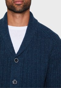 Donkerblauwe gebreide cardigan met een sjaalkraag, voorzien van een geribbelde textuur en twee grote zwarte knopen aan de voorkant.