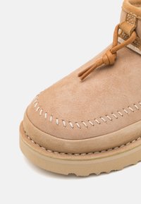 Tan suede ankle boot featuring a rounded toe, contrasting white stitching, and a braided leather lace tie. Flat rubber sole with textured detail.